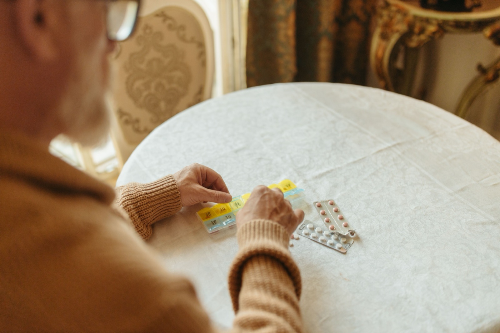 An older man's hands sorting pills into a weekly organizer on a table.