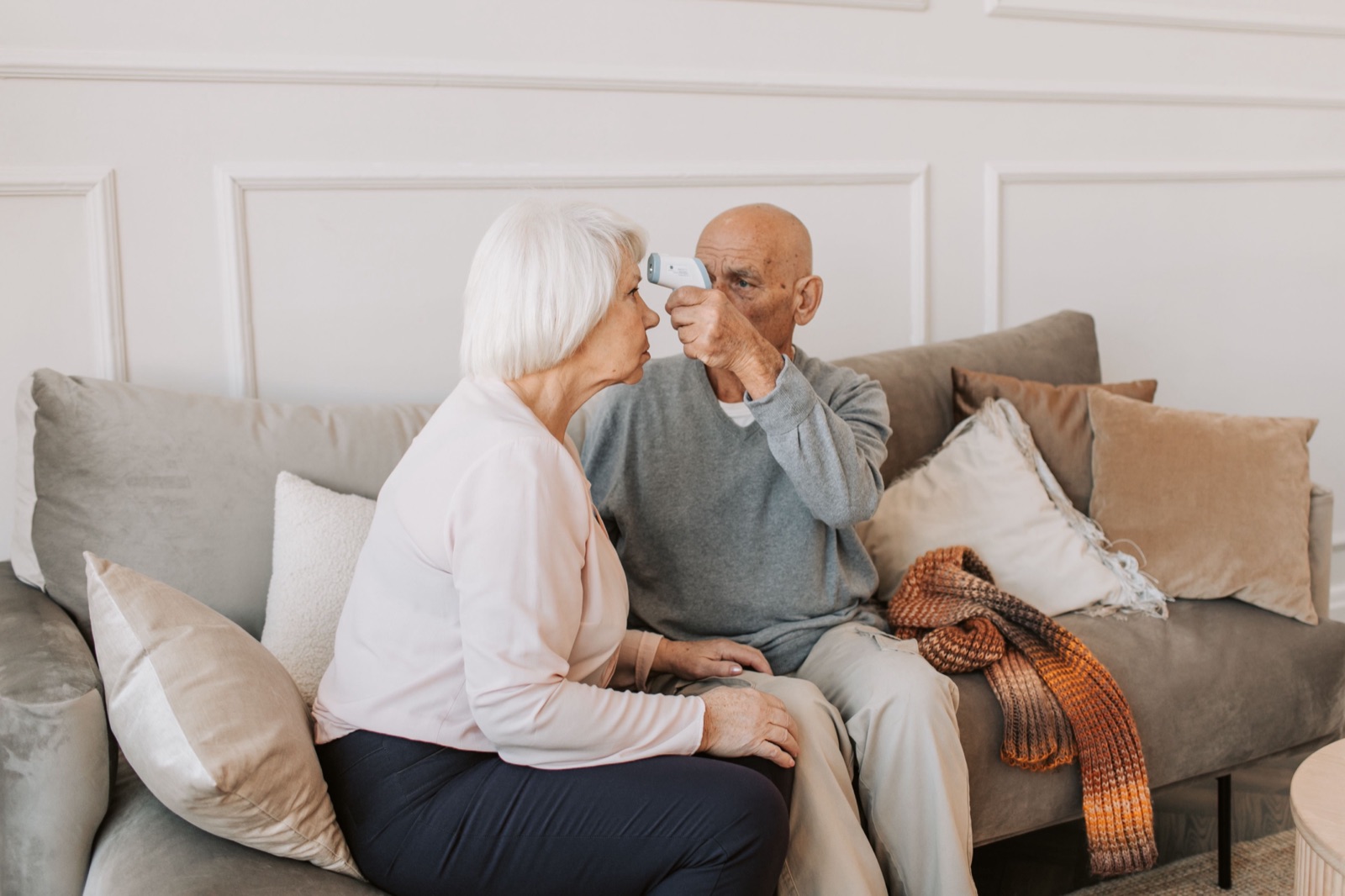 An older couple on their couch at home as he checks his temperature.
