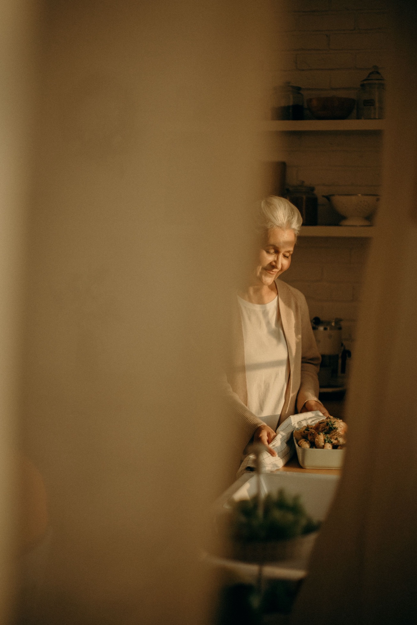 An older woman smiling as she prepares food in her kitchen in warm evening light.